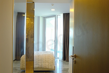 Image of a furnished bedroom. The room features a double bed, a headboard, bedside tables, a wardrobe, and modern wall sconce lighting. White walls and wooden parquet flooring contribute to a spacious and bright feel in the room.