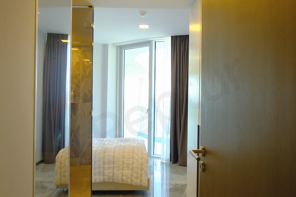 Image of a furnished bedroom. The room features a double bed, a headboard, bedside tables, a wardrobe, and modern wall sconce lighting. White walls and wooden parquet flooring contribute to a spacious and bright feel in the room.