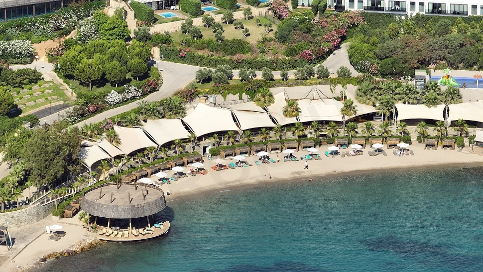 A sunny view of a private residence beach in Bodrum, with clear turquoise sea and fine sand. Sun loungers and umbrellas are neatly arranged on the beach.