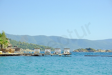 A private wooden pier extending over the calm blue sea under sunny skies. The pier is bathed in sunlight, surrounded by clear, azure water.