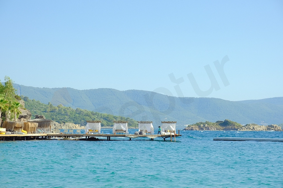 A private wooden pier extending over the calm blue sea under sunny skies. The pier is bathed in sunlight, surrounded by clear, azure water.