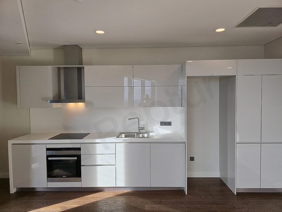 Bright apartment kitchen with modern white cabinets, wooden countertop and built-in appliances.