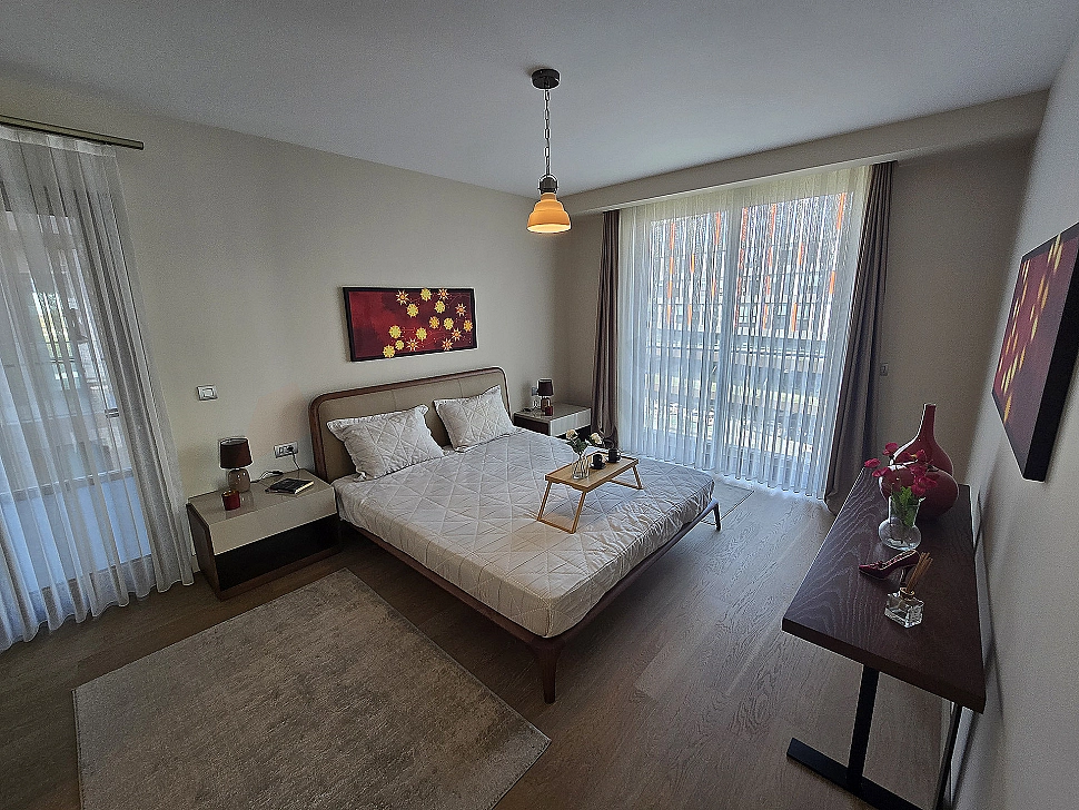 Modern bedroom interior: double bed, nightstands, lamps, rug, and window with city view.